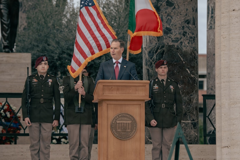 A man in a suit speaks behind a lectern as four people in military dress uniforms stand behind him. One of them in holding the American flag while another holds the Italian flag.