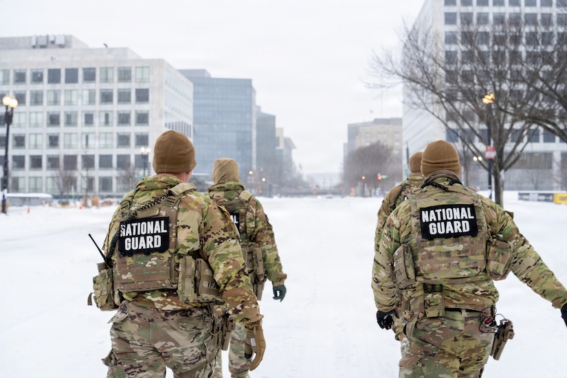 Four people in camouflage military uniforms walk along a snow-covered city street.