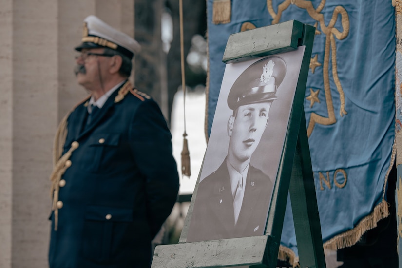 A portrait of a man in a military dress uniform is displayed on an easel during a commemoration ceremony in Italy. An elderly man in a military dress uniform stands in the background.