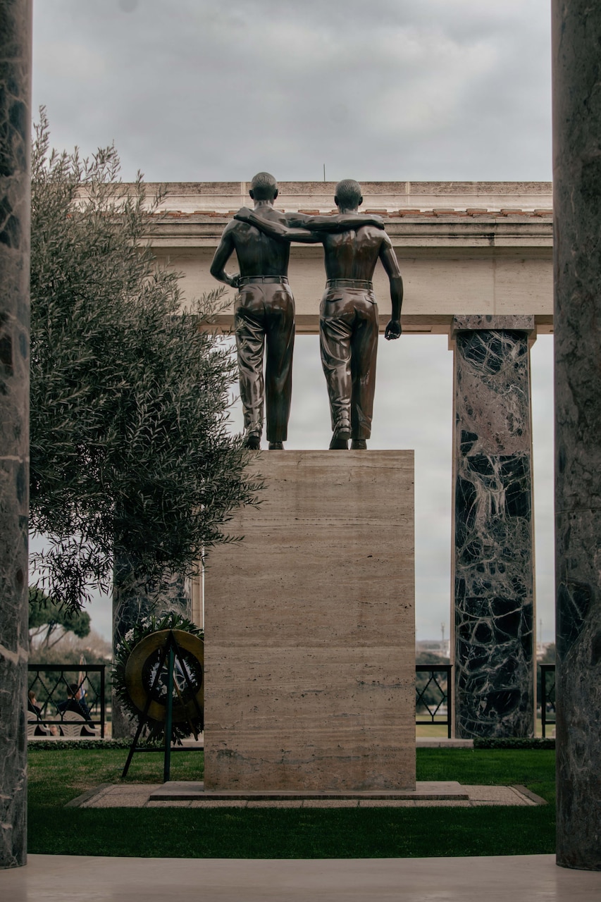 The back of a monument of two men with their arms over each other's shoulders is displayed during a commemoration ceremony in Italy. There is a wreath displayed on a stand in front of the monument.