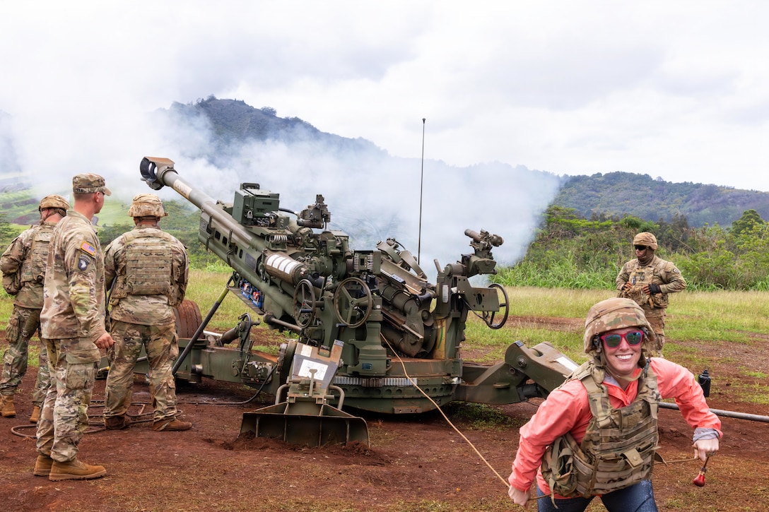A woman wearing a camouflage military vest and helmet smiles and runs, pulling a cord away from a cannon-like weapon, which explodes in the background. Four soldiers in similar attire stand near the weapon system.