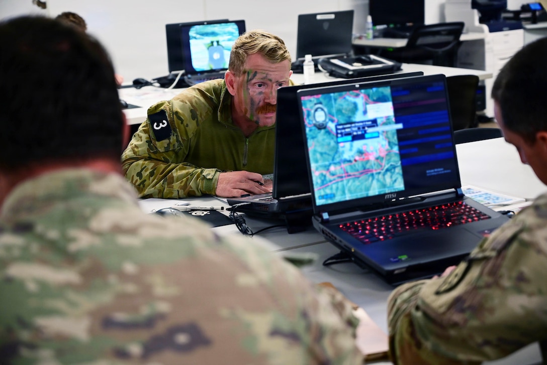 A man sitting at a table wearing a camouflage military uniform and face paint stares at a laptop; two soldiers in similar attire sit on the opposite side, one looking at a laptop screen coordinate. Similar laptops are in the background.