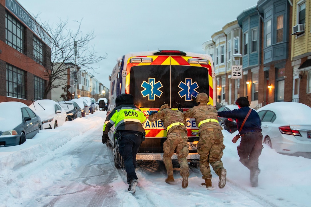 Two people wearing camouflage military uniforms and two people in civilian and fire department attire push an emergency medical vehicle down the street.