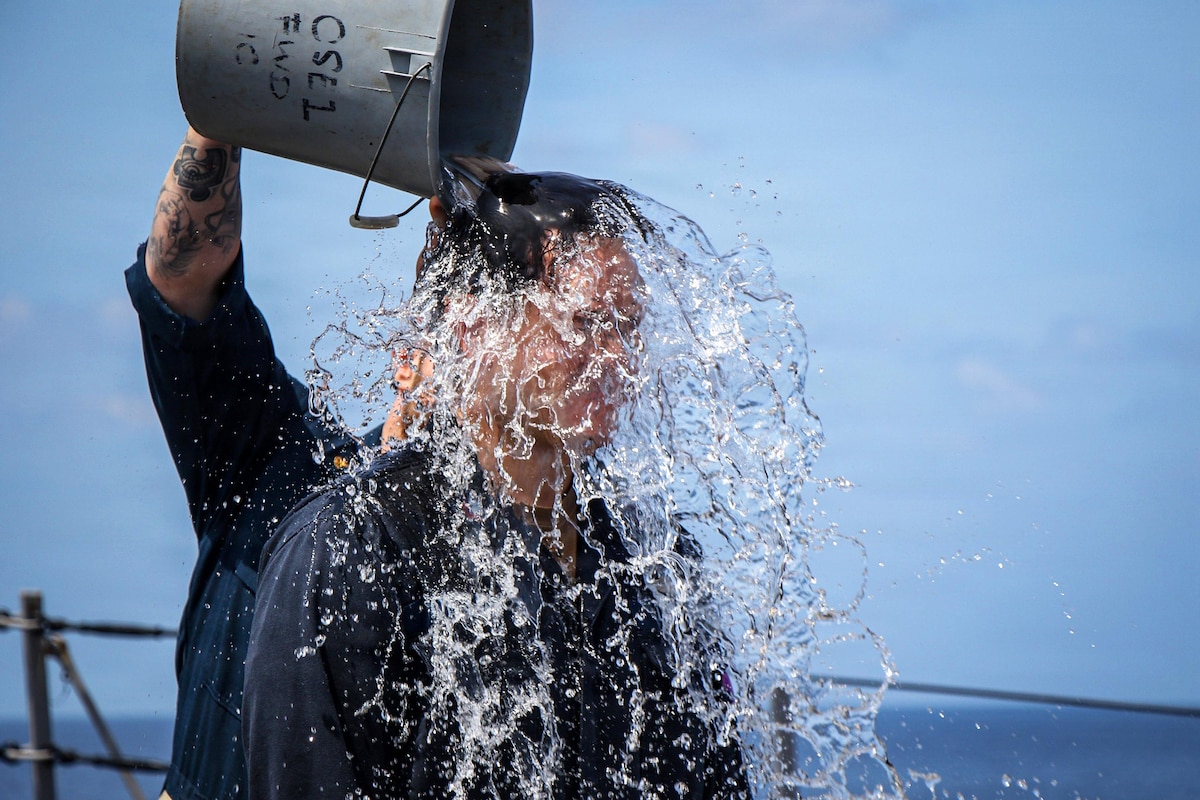 A sailor in casual attire dumps a bucket of water on the head of another sailor wearing similar attire outside under a blue sky.