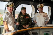 Two naval officers stand and one man sits on the bridge of a ship