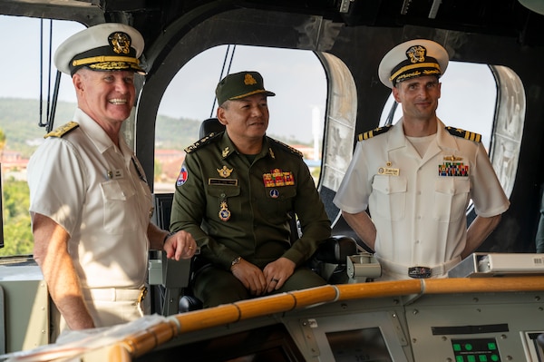 Two naval officers stand and one man sits on the bridge of a ship