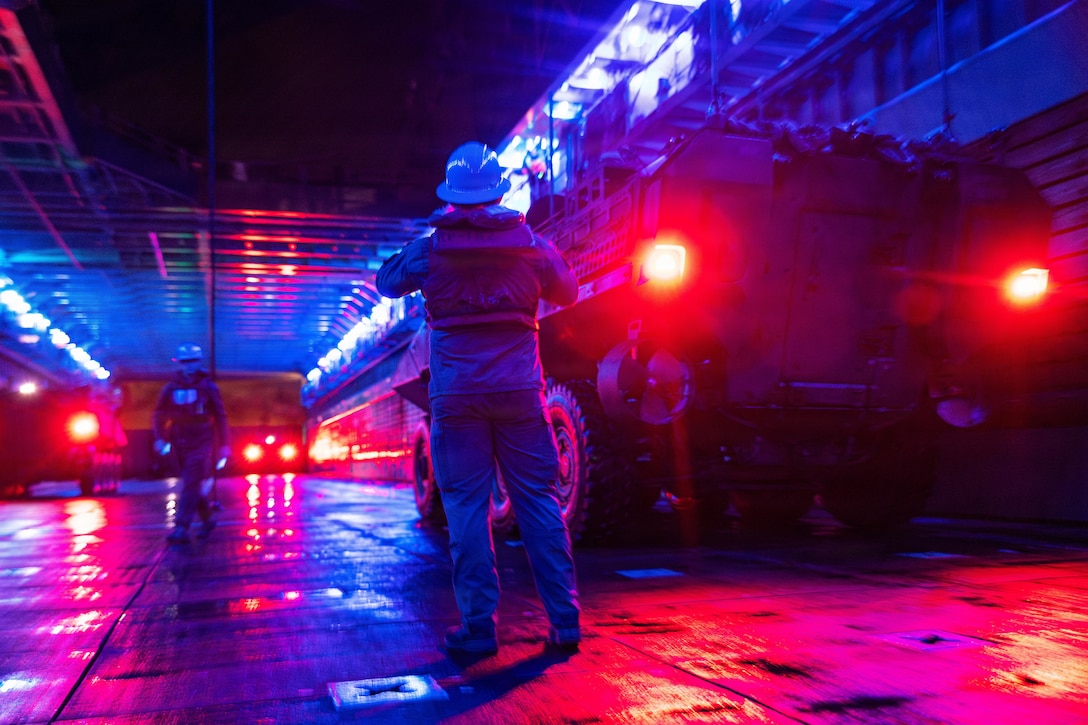 A person wearing safety clothing and a hard hat directs a vehicle at night with glaring red lights; a person wearing similar attire is walking toward the person; several other vehicles with glaring red lights are in the background.
