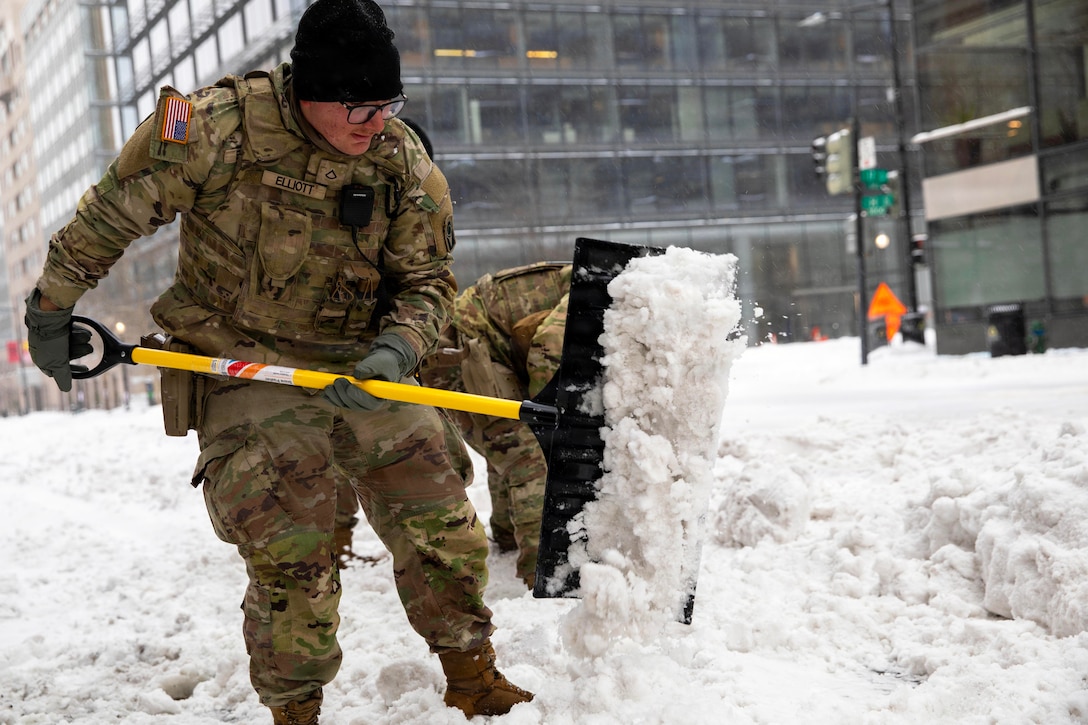 A man wearing a camouflage military uniform shovels snow in a city, with another person wearing similar attire in the background.