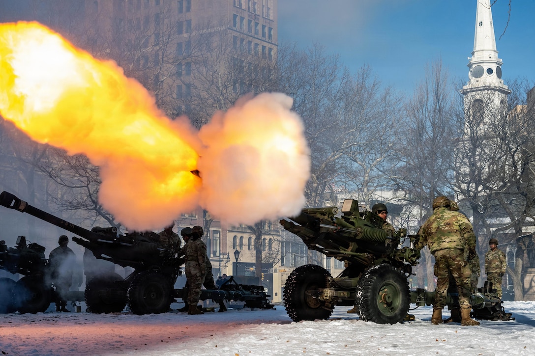 A large explosion of bright yellow and white smoke is released from a cannon, as several people wearing camouflage military uniforms stand behind it. Two other cannons and several people in similar attire are positioned on the left.