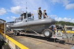 Two people in uniform stand atop a rigid hull inflatable boat that's being wheeled in on a dock during daylight.