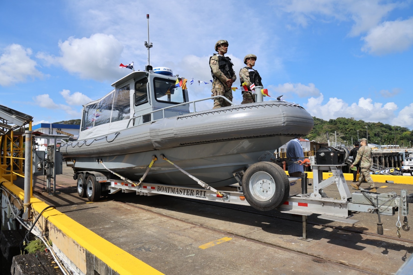 Two people in uniform stand atop a rigid hull inflatable boat that's being wheeled in on a dock during daylight.