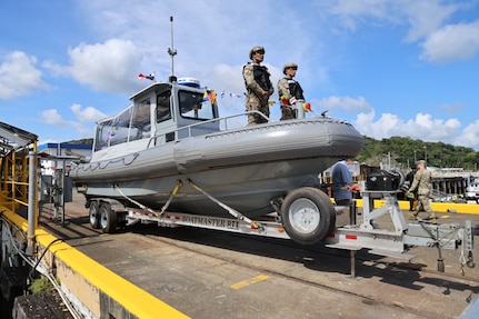 Two people in uniform stand atop a rigid hull inflatable boat that's being wheeled in on a dock during daylight.