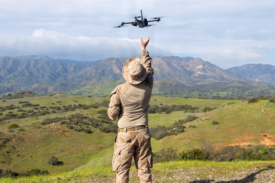 A person stands on a hillside wearing a camouflage military uniform and reaches for a drone hovering during daytime, with clouds overhead and mountains in the background.