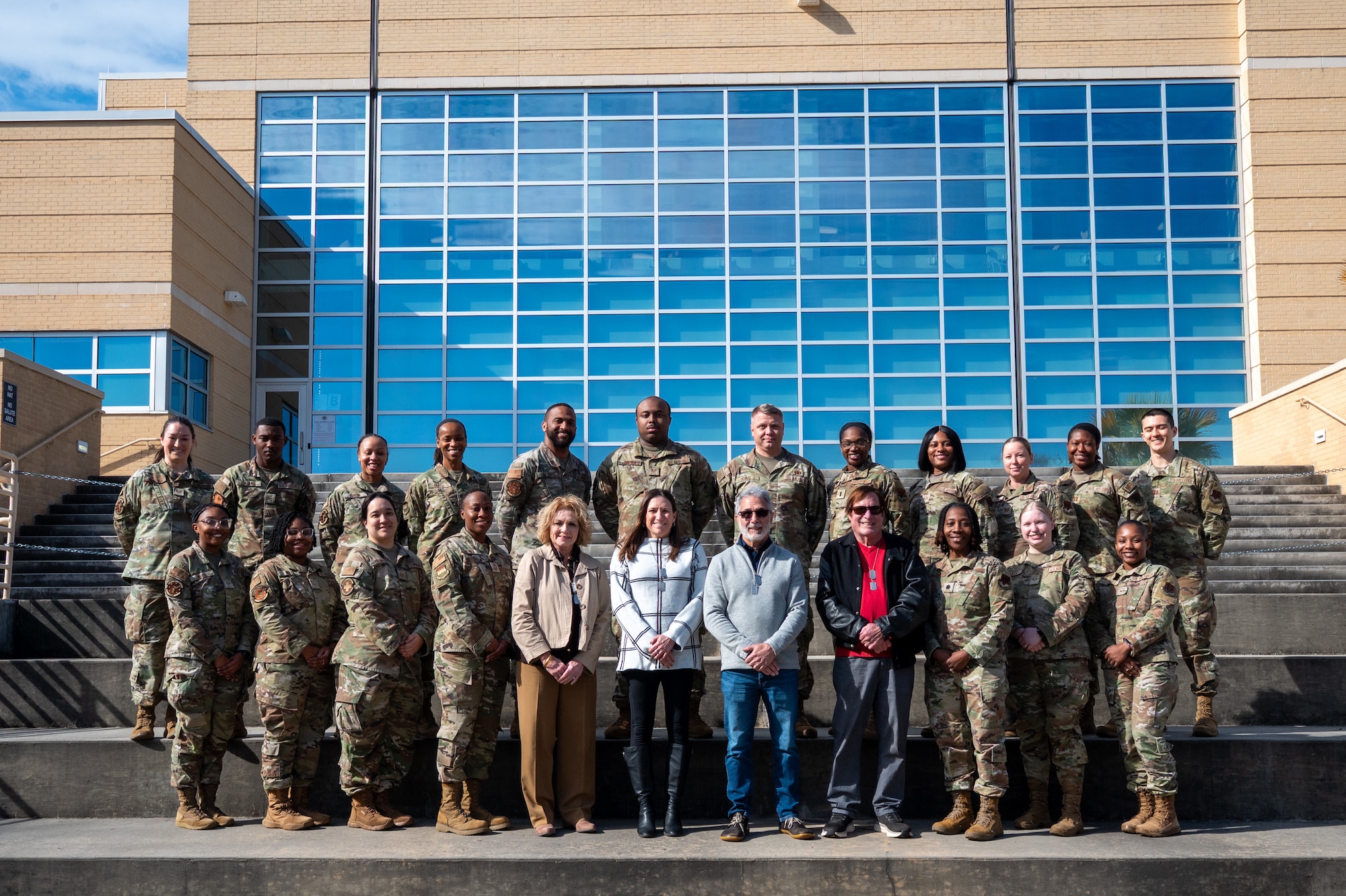 Four civilians and multiple U.S. Air Force Airmen pose for a group photo on steps outside of a medical facility