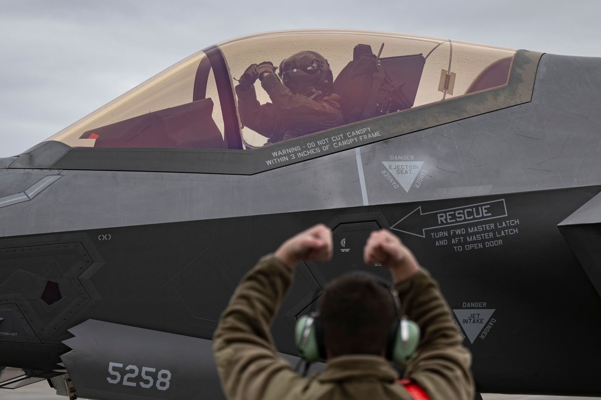 A photo of an F-35 pilot in a cockpit signaling to a maintainer on the ground.