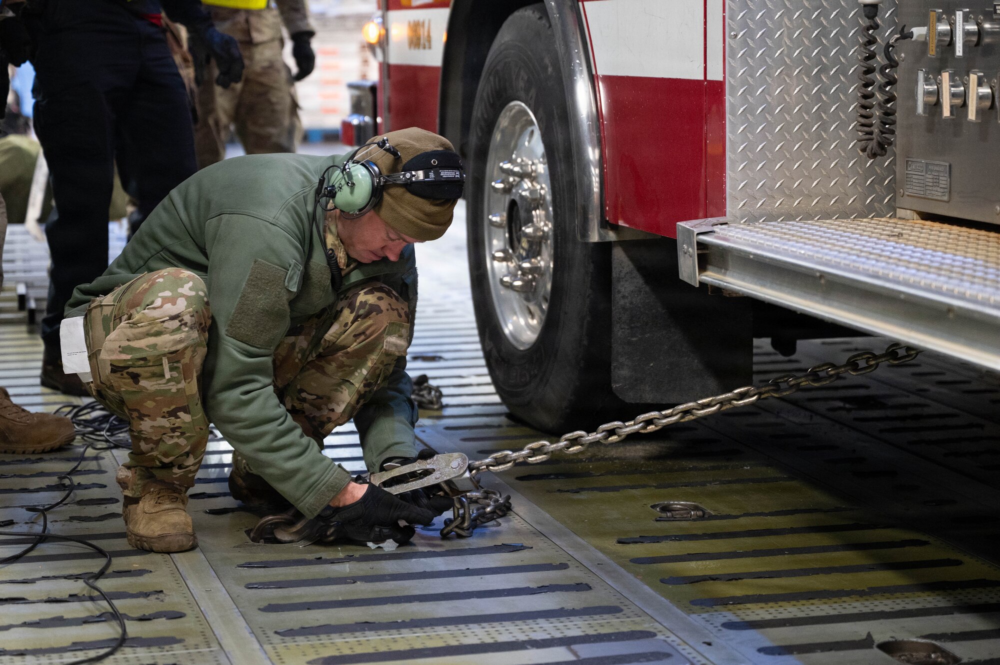 An Airman crouches on the ground next to the wheel of a firetruck, attaching a chain to the floor of a C-17 aircraft to secure the vehicle.