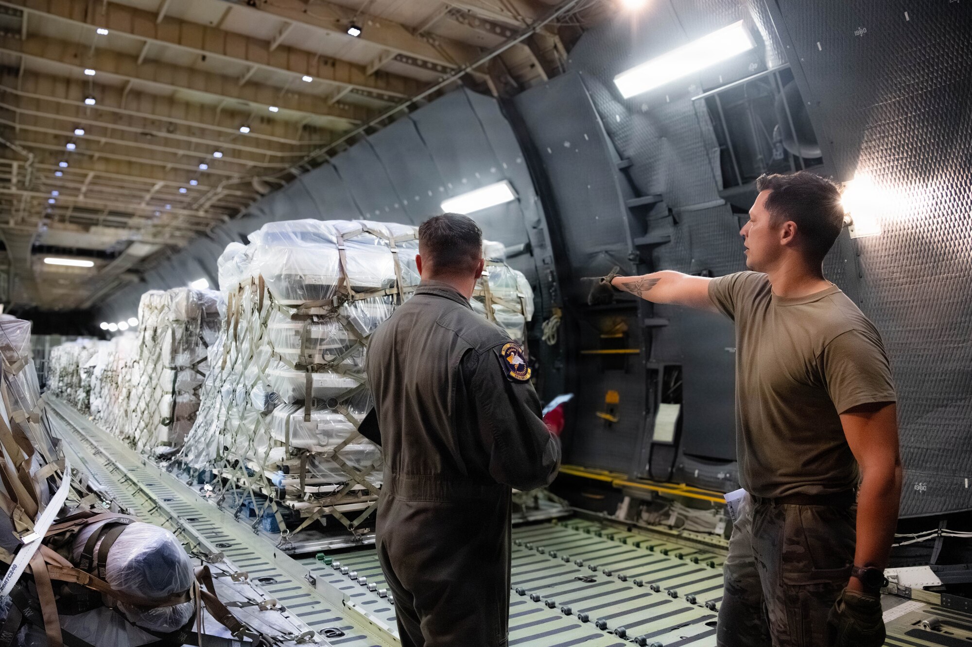 Two Airmen speak to each other, one points towards a pallet of cargo in the background.
