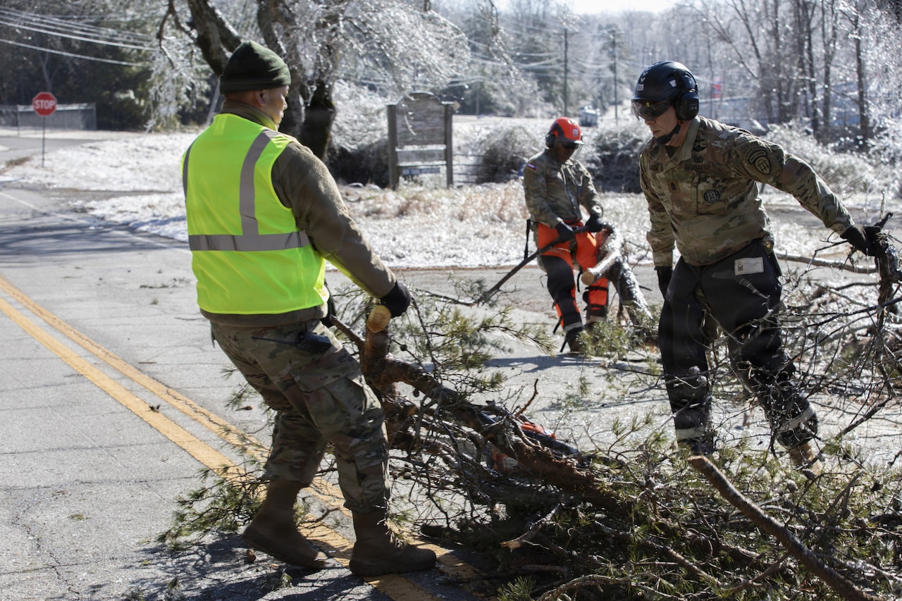 Three men dressed in camouflage military uniforms pull tree branch debris away from a road.