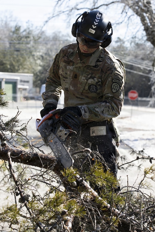 A man dressed in a camouflage military uniform uses a hand saw to cut a tree branch.