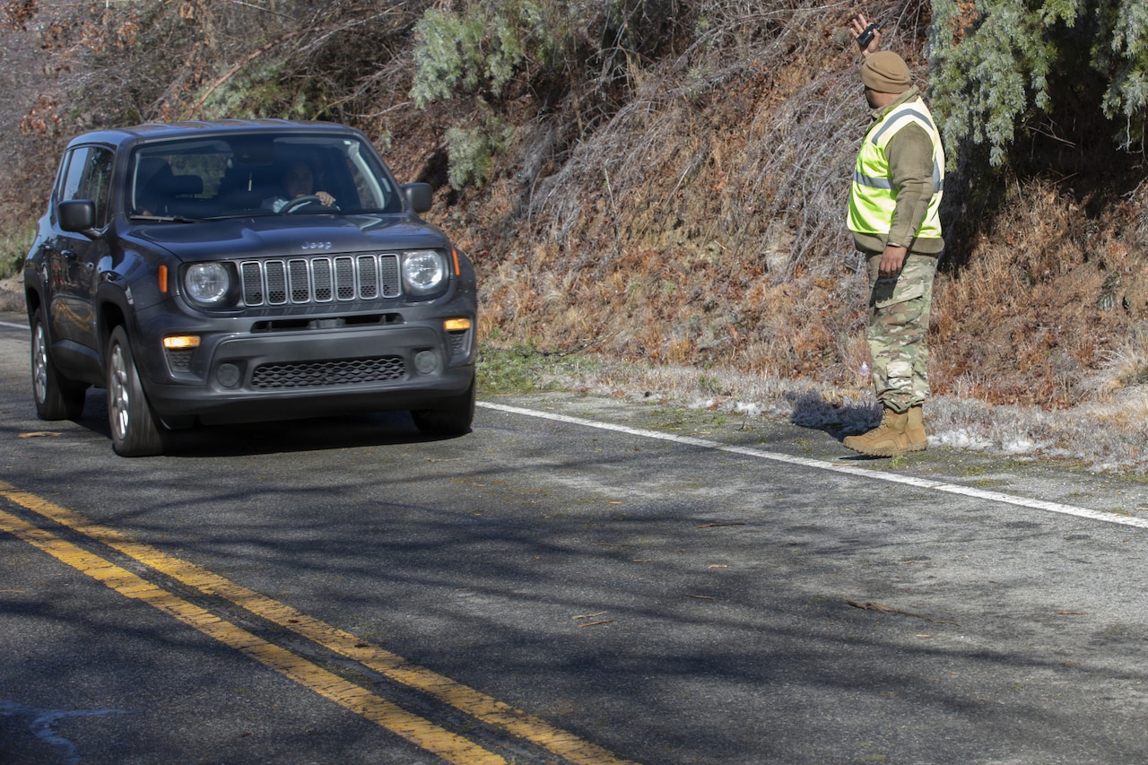 A man wearing a hazard vest and camouflage military uniform stands by a roadside to direct a car away from a work zone.