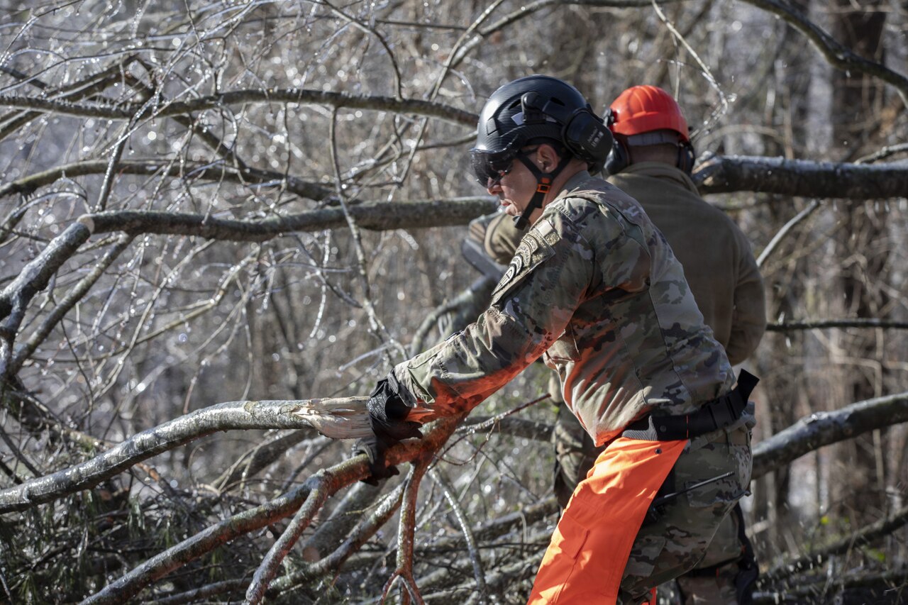 Two men dressed in camouflage military uniforms remove broken branches.