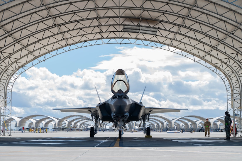 Two people in camouflage military uniforms stand next to a large military jet parked on the tarmac under an aircraft tent. The background is filled with more tents and aircraft. The cockpit hatch is open on the jet.