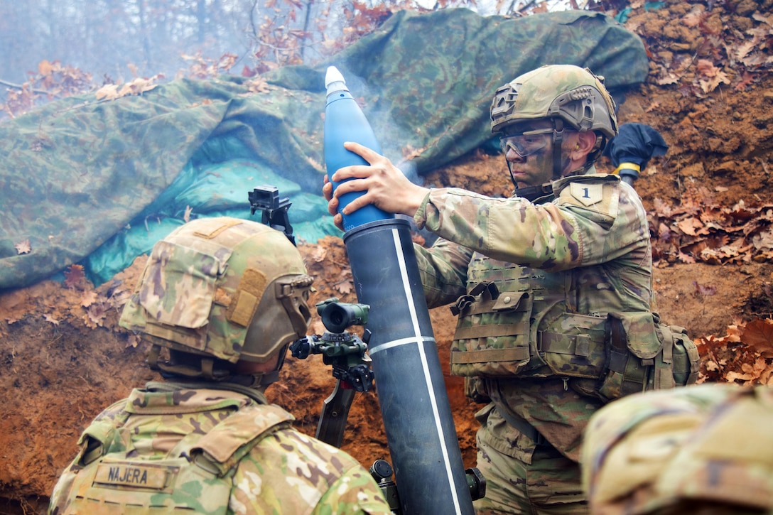 A man standing on a rocky, muddy hill, wearing a camouflage military uniform and helmet, places a rocket into a cylinder as a man in similar clothing looks on.