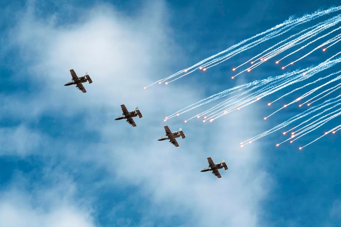 Four planes fly across a blue sky with small yellow flares and white smoke trails behind them.