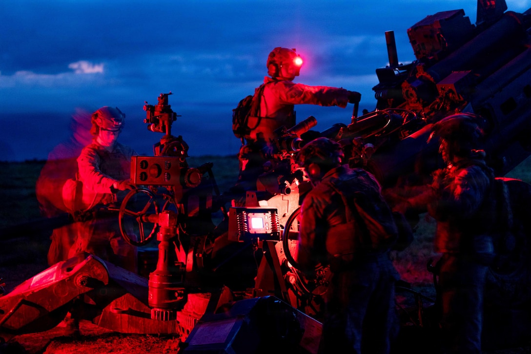 Several people wearing camouflage military uniforms and helmets position gears on a large gun at night under a cloudy sky; one person has a glowing red light on the helmet.
