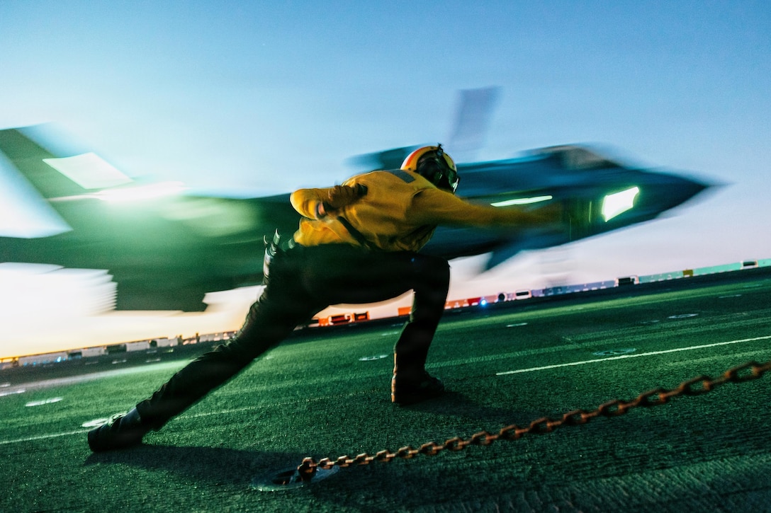 A person in yellow and black attire and headgear signals to a pilot taking off in a jet from a ship's flight deck.