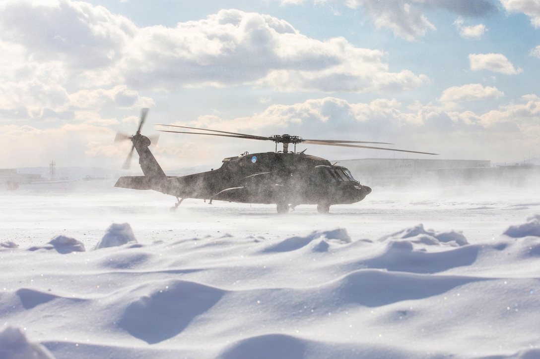 A helicopter lands in the deep snow on a clear day, with several buildings and equipment in the background.