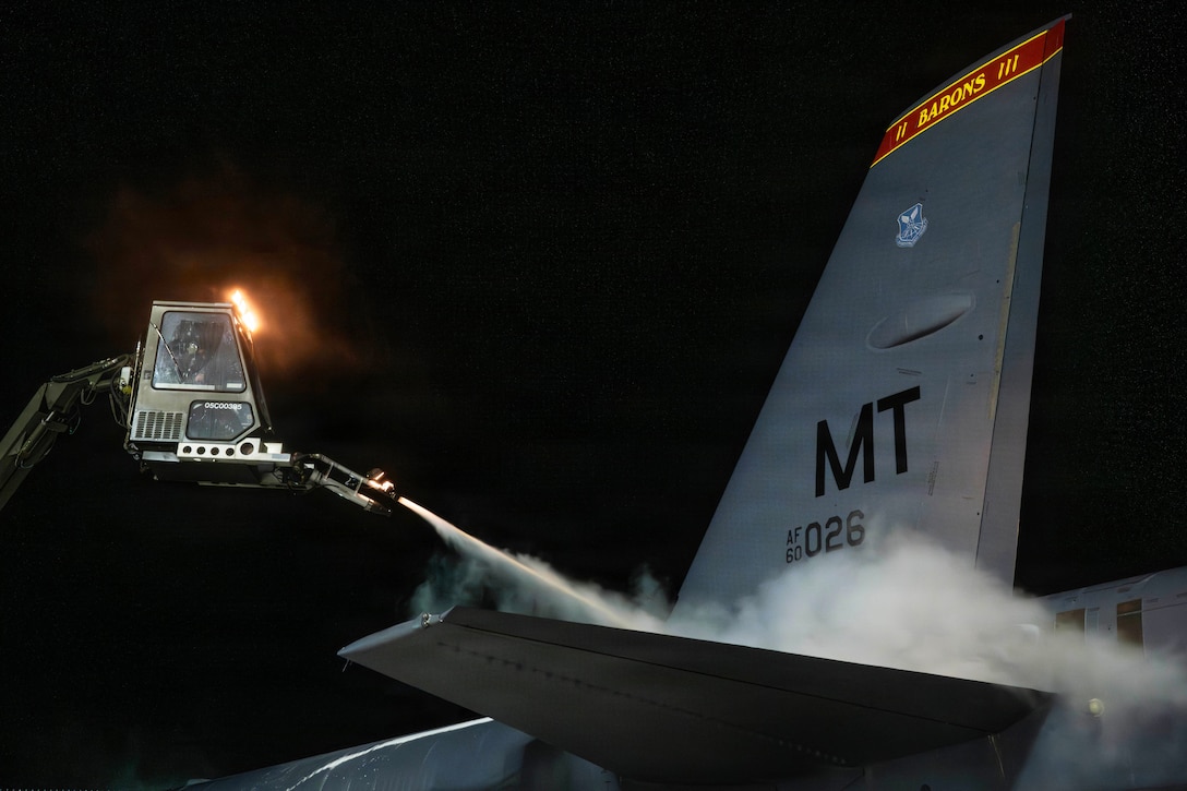 A person in a crane hovers over the rear of a military aircraft at night to blow deicing solution onto the wing, while white smoke-like plumes rise into the air as the solution makes contact with the plane.