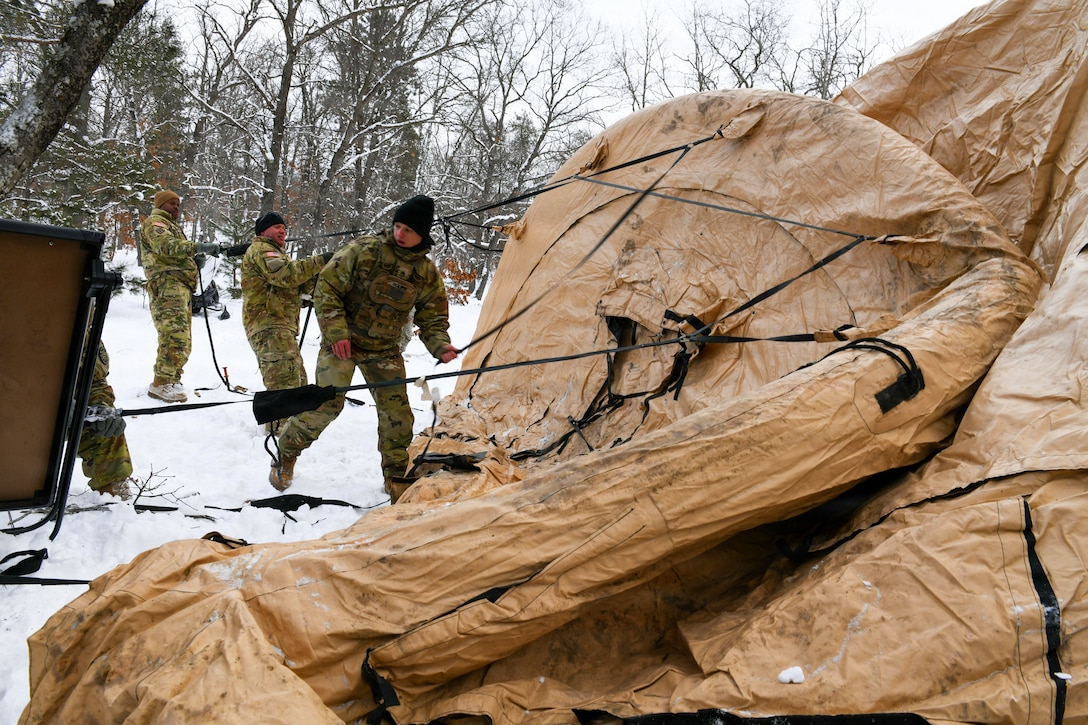 Three people standing in a row outside in the snow, wearing cold-weather camouflage military uniforms, pull cords to set up a tent, with snow-covered trees in the background.