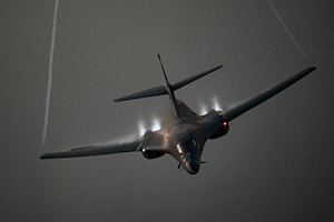A large military aircraft flies through the air with a trail of water vapor coming off the wings.