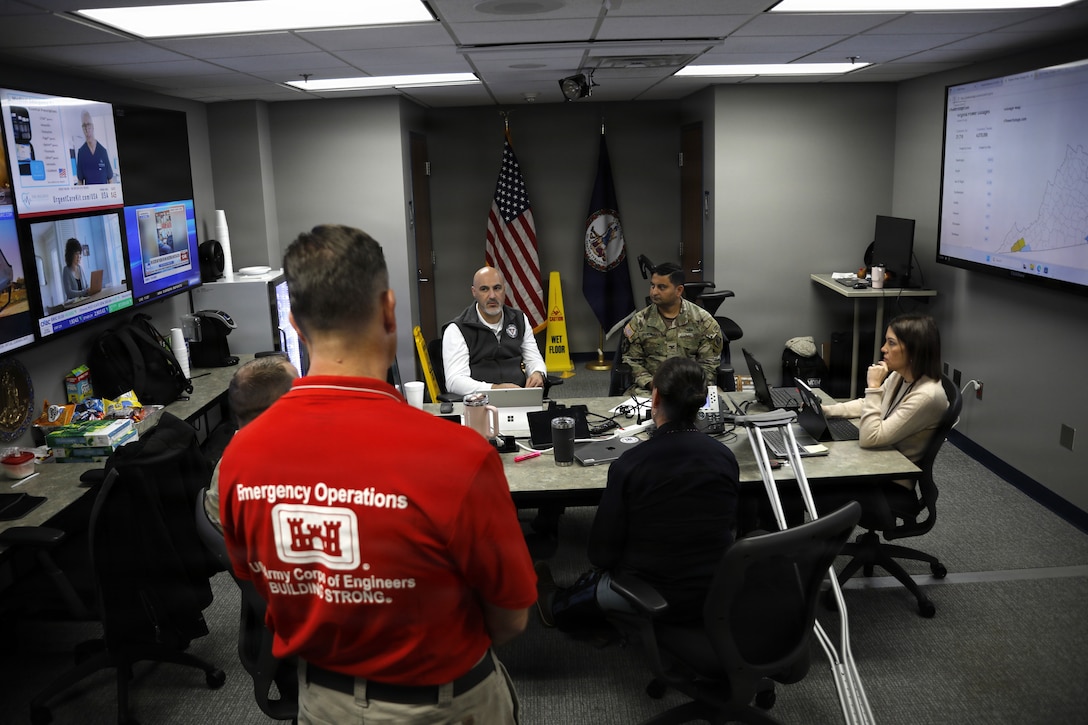 A group of people sit in a small conference room, there are multiple tv screens on one wall and a white board on another. One gentlemen in a red emergency operations shirt is seen standing closest to the camera.