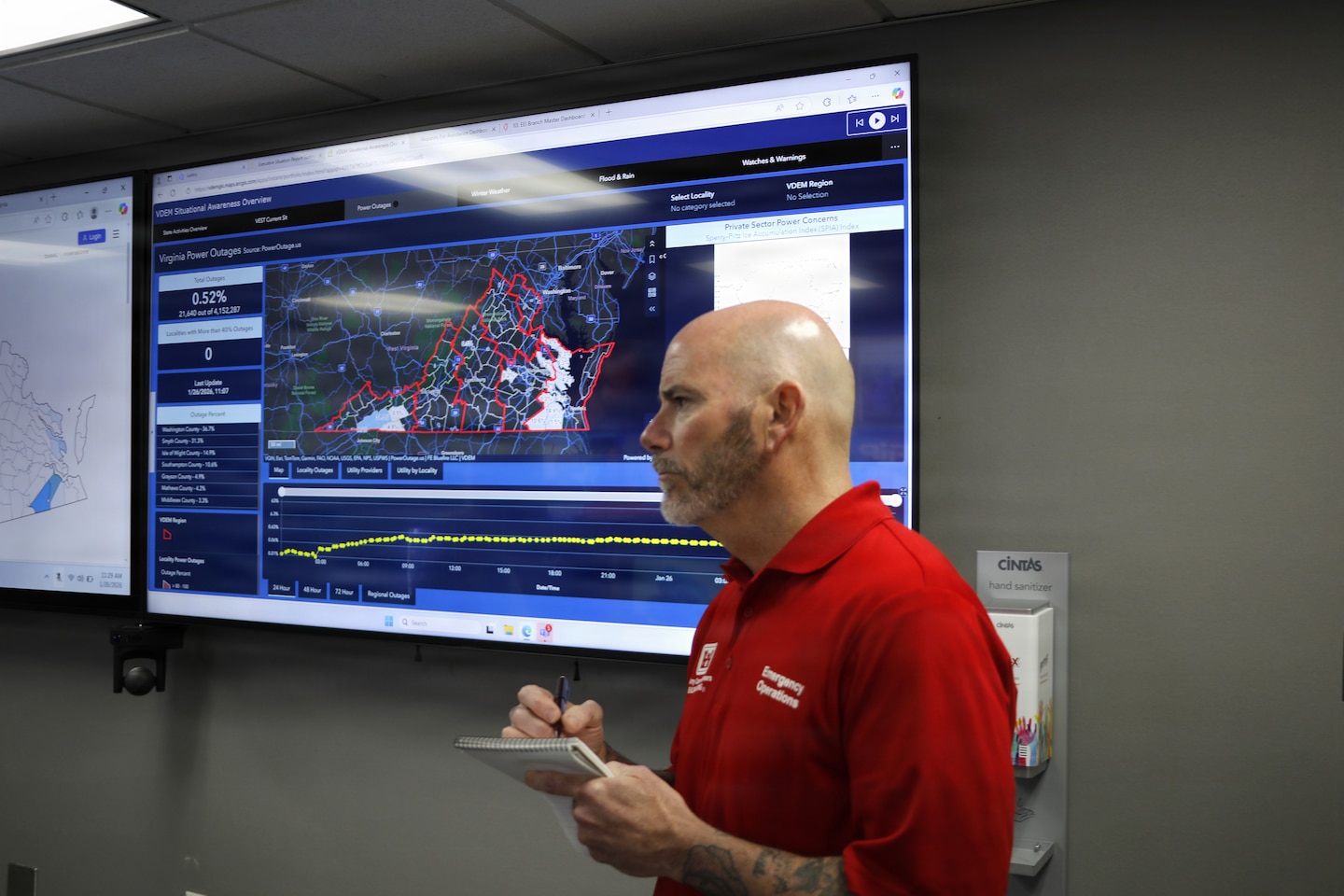 A man is seen wearing a read collard shirt standing in front of a large tv monitor with the state of Virginia on it.