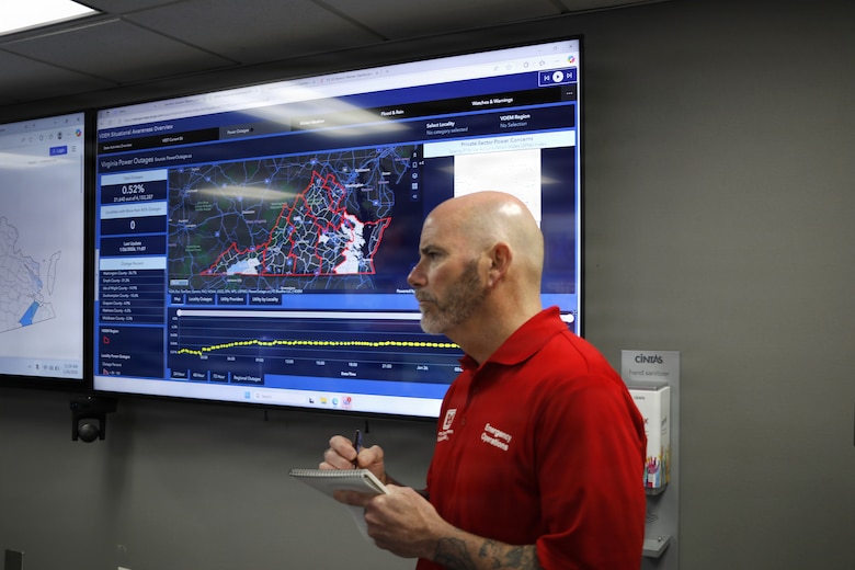 A man is seen wearing a read collard shirt standing in front of a large tv monitor with the state of Virginia on it.