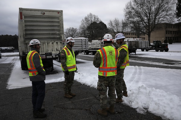Soldiers are seen standing in a group wearing hard hats and protective vests. There are generators on flatbeds in the background, and snow and ice is covering the ground.