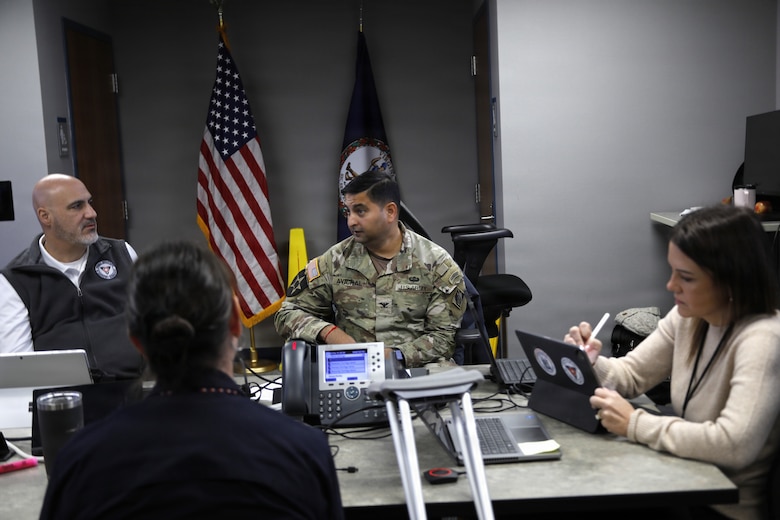a group of men and women are seen sitting around a small conference room table. One man is a Soldier.