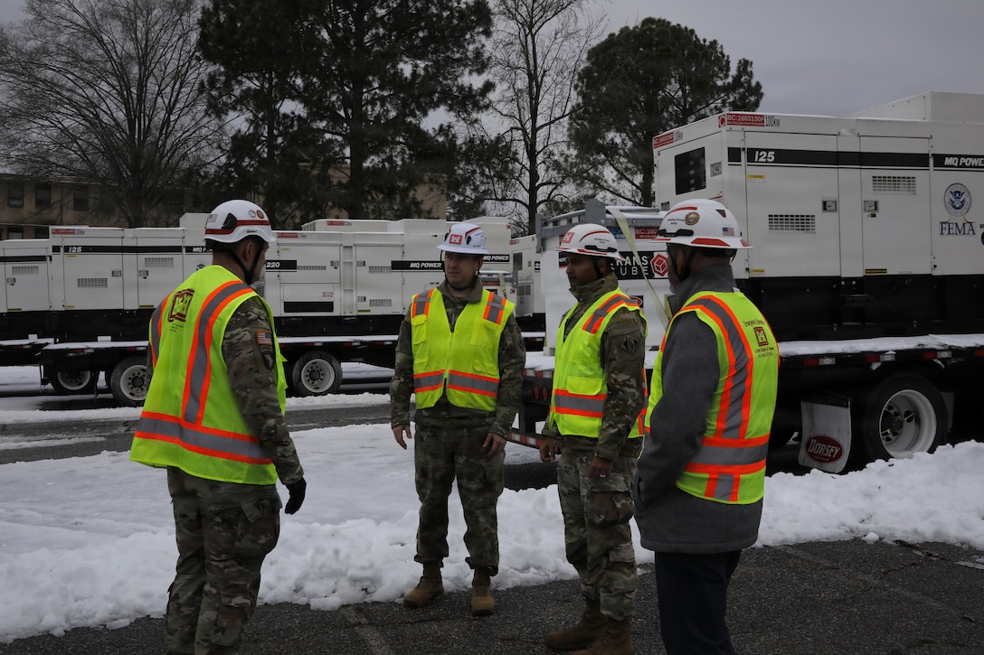 Soldiers in Hard Hats and protective vests are seen talking to each other with generators sit on flatbeds in the background, the ground is covered in snow and ice.