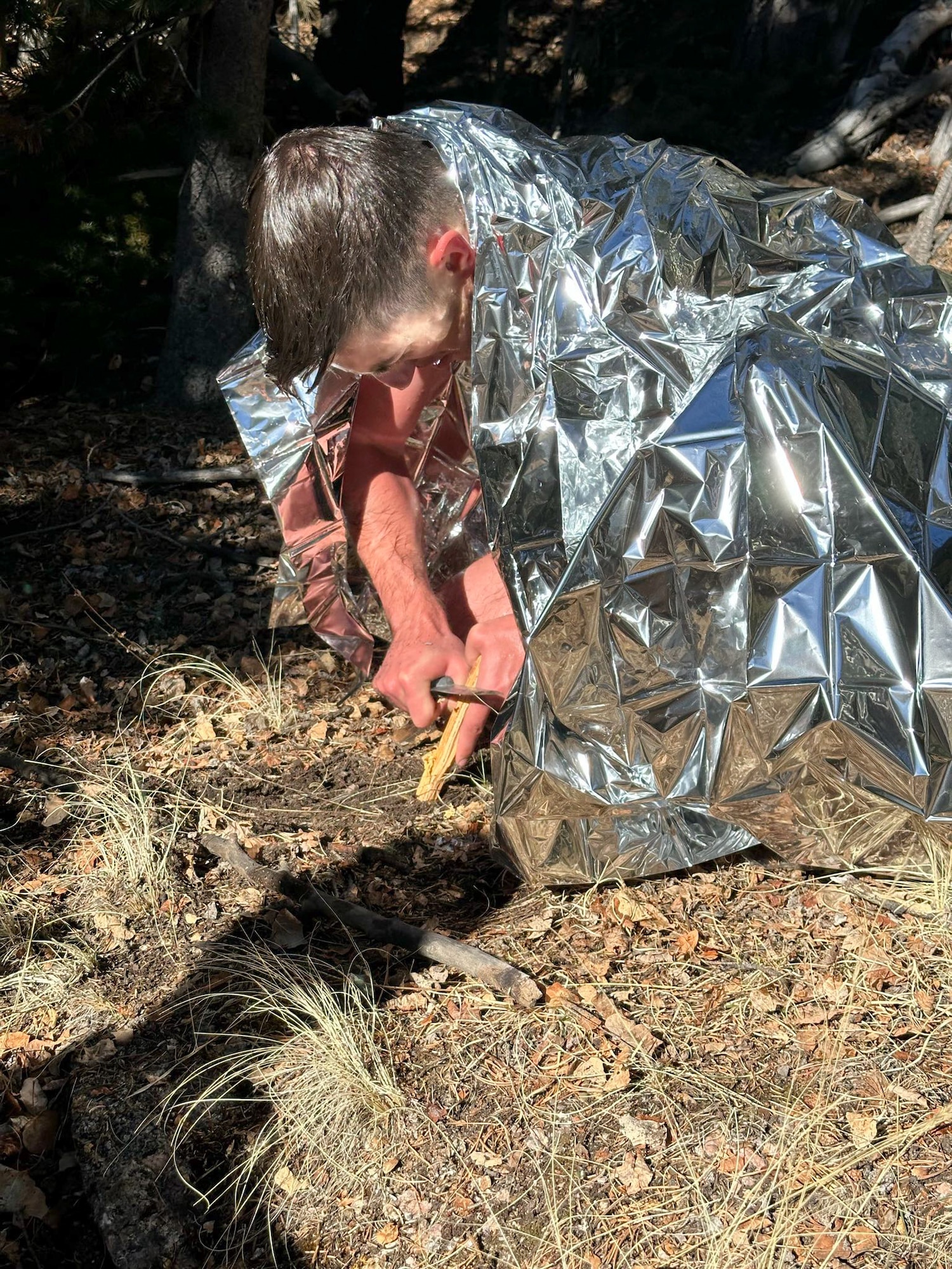 Humanic is using an emergency blanket to conserve heat while attempting to build a fire.