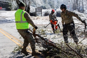 Three men wearing camouflage military uniforms pull tree limbs across a road in snowy conditions.