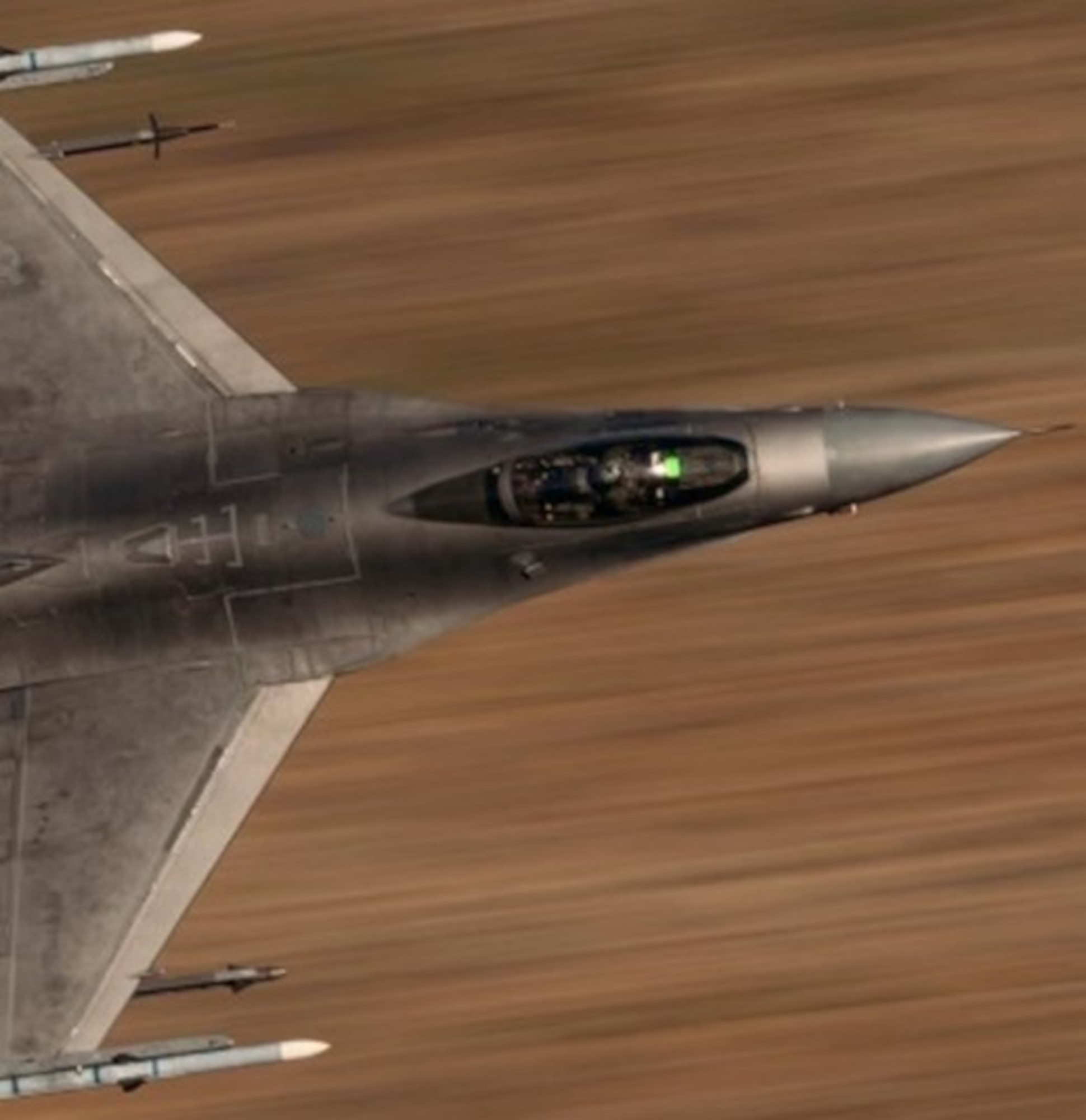 A headshot of an F-16 Fighting Falcon flies with a blurred desert landscape background.