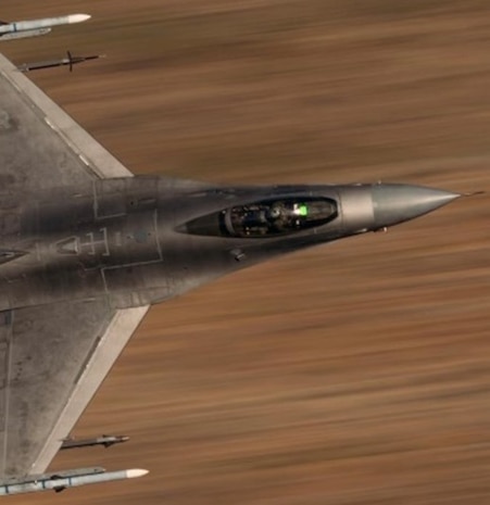 A headshot of an F-16 Fighting Falcon flies with a blurred desert landscape background.