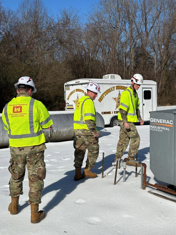 Three soldiers inspect a large electrical connection point in an area that is covered in snow and ice.