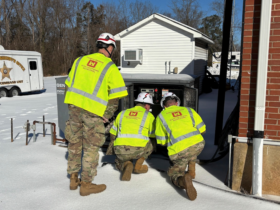 Three soldiers inspect a large electrical connection point in an area that is covered in snow and ice.