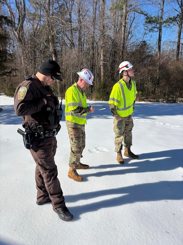 Two soldiers stand with a Sheriffs Deputy looks in an area that is covered in snow and ice.