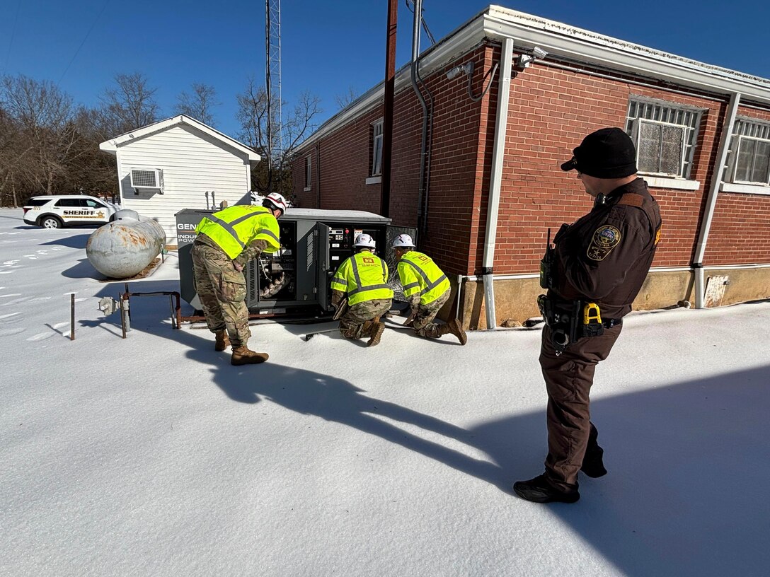 Three soldiers huddle around an electrical connection point as a Sheriffs Deputy looks on. The area is covered in snow and ice.