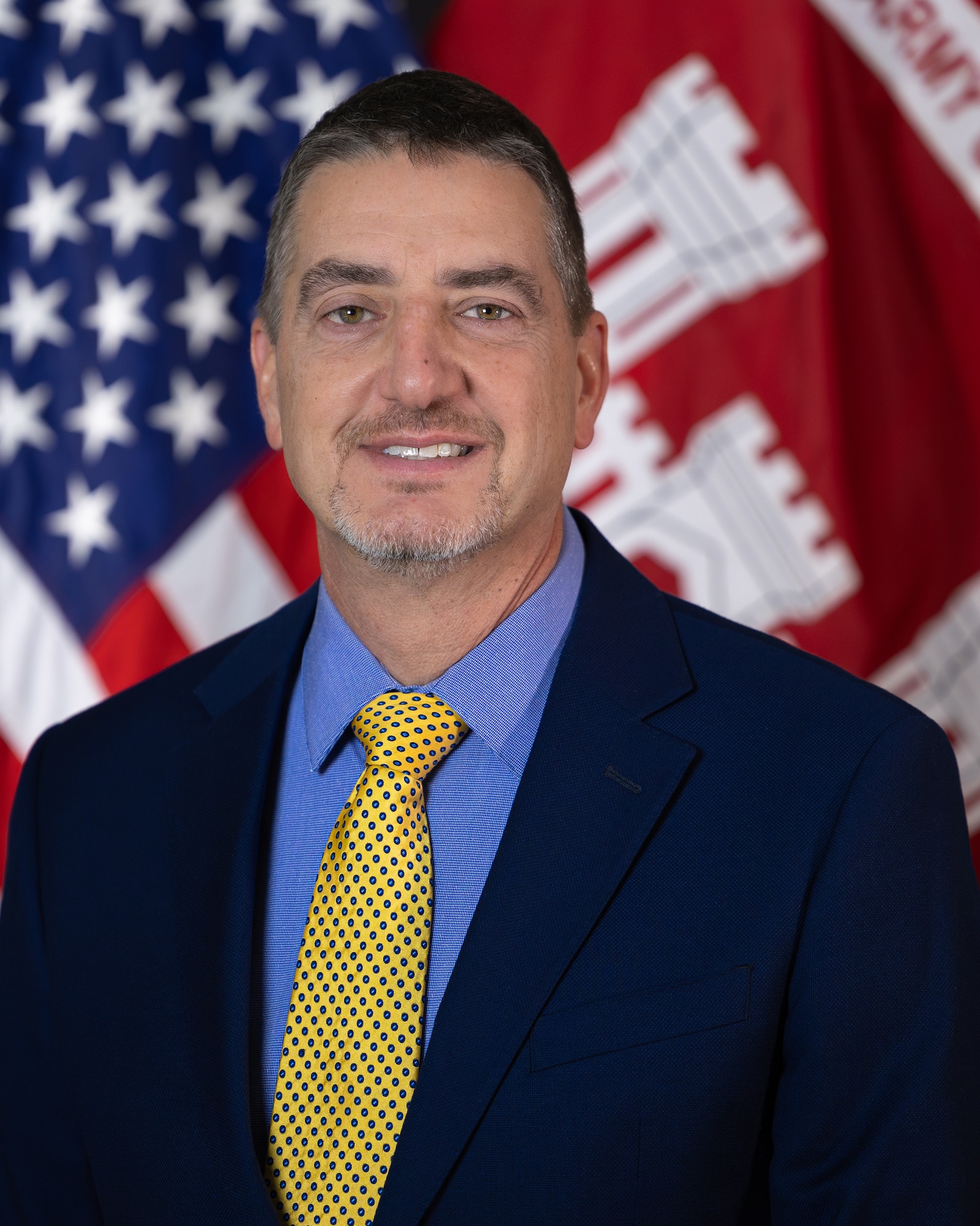 A man in a black suit, blue shirt, and yellow tie smiles in front of an American flag and a red and white flag.