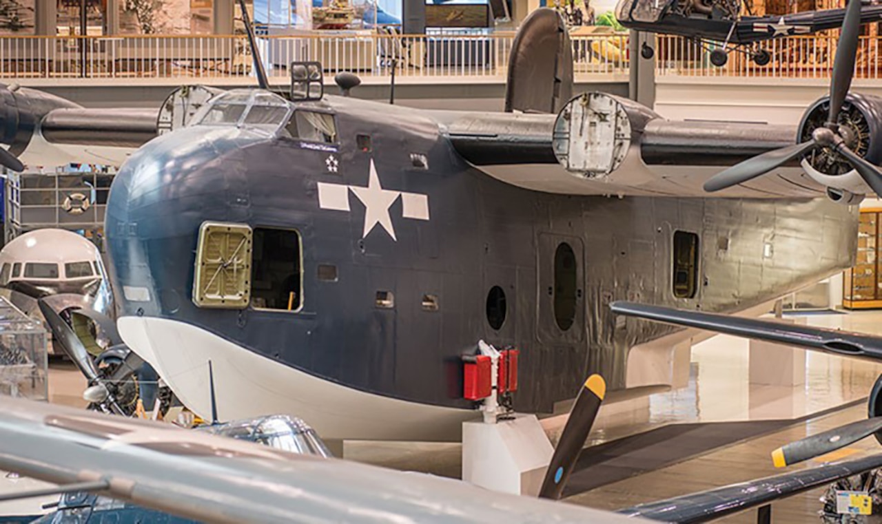 A large seaplane sits on the tiled floor of a museum. Other aircraft and their wings can be seen nearby.
